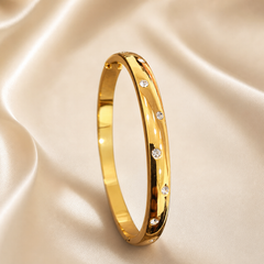 A single rigid oval bangle of yellow gold with a high-polish mirror finish, formed as a continuous solid band of medium gauge with a softly domed outer profile and flat inner surface. Set into the outer face are round, clear, colourless faceted stones in flush (gypsy) settings, each sitting level with the metal surface in its own small circular seat; the stones are of similar small size and are distributed at irregular, uneven intervals around the visible face rather than in an even grid, with some