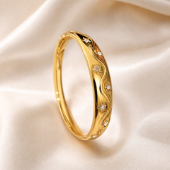 A single rigid bangle (kada) in polished yellow gold with a bright mirror finish. The band is a continuous closed oval of moderate width, slightly tapered, with a plain polished inner surface. Around the outer face runs an engraved relief pattern of flowing vines and floral motifs.
