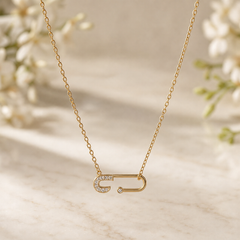 One necklace in yellow-gold-toned metal with a polished finish. A single fine-gauge oval-link cable chain forms one continuous strand, its two ends joined by small jump rings to the ends of a single fixed safety-pin pendant that hangs horizontally as the central feature; the chain run measures roughly 18 times the pendant's width, and no extender is present.