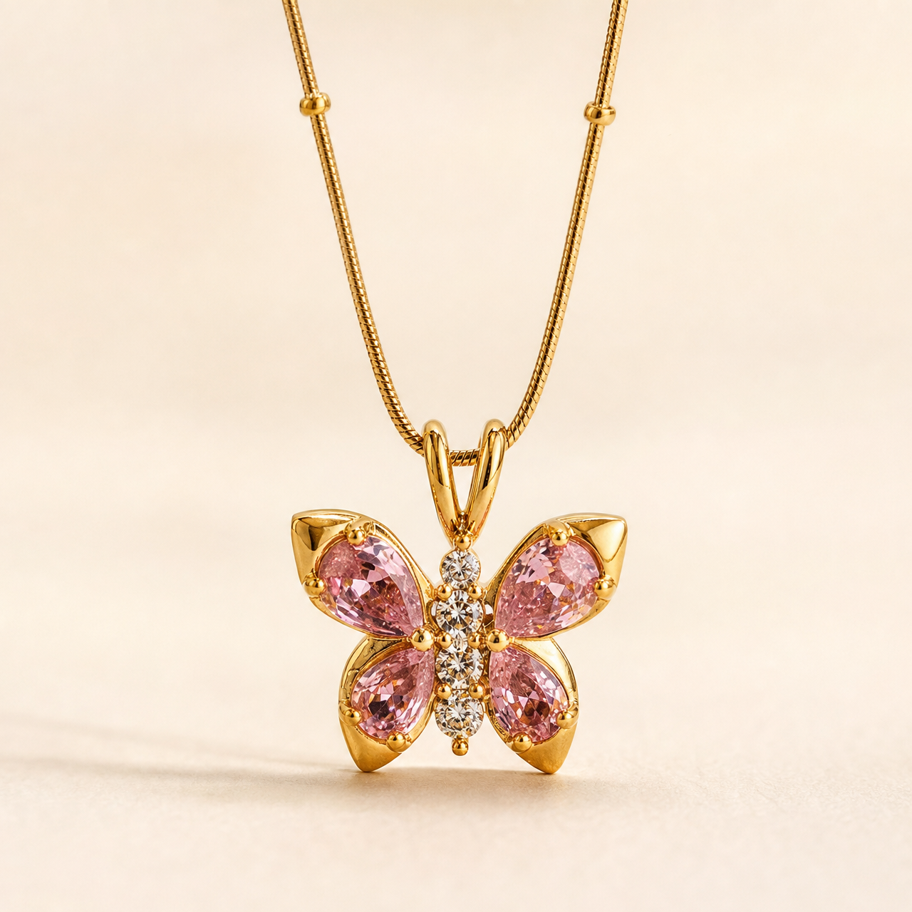 No earrings are present in this image. The visible product is a single pendant necklace: a yellow-gold-tone fine snake chain with small spaced bead stations, suspending a butterfly-shaped pendant through a double-loop bail. The pendant has polished gold-tone metal with four pear-cut pink faceted stones forming the wings in prong settings, and a vertical body of small round clear faceted stones. Pendant drop is roughly one and a half times its widest span.
