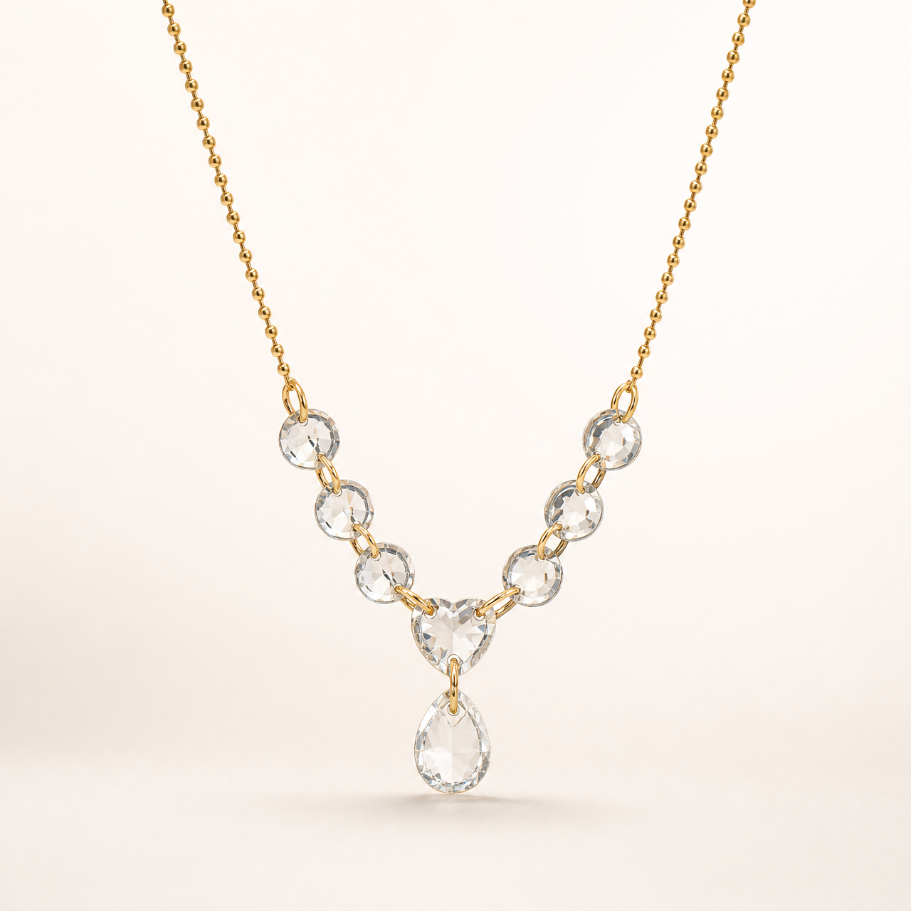No earrings are present in this photograph; the visible item is a single necklace. It has a fine gold-toned beaded chain of yellow-metal colour with polished finish. The chain descends to a small pendant cluster: a row of small round clear faceted elements, each drilled and linked by jump rings, converging on one larger clear heart-shaped faceted element, from which a single larger clear teardrop-shaped faceted element dangles, jump-ring mounted.