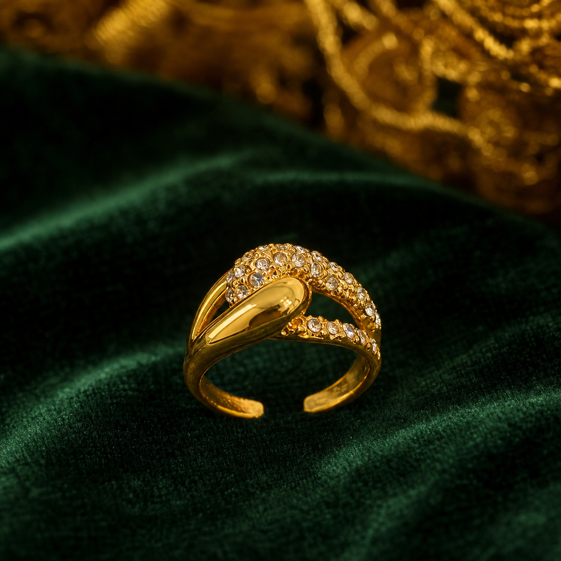The photograph shows one ring, not earrings: a single gold-tone finger ring with a polished yellow-metal finish. The band splits into two curving arms that cross over one another; one arm is a smooth, polished solid-metal ribbon, while the other carries a continuous pavé field of small round clear faceted elements set into the metal. The band is open-backed with an adjustable gap. Construction is a single rigid piece with no articulation, hinges, drops or dangling components.