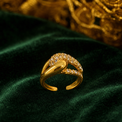 The photograph shows one ring, not earrings: a single gold-tone finger ring with a polished yellow-metal finish. The band splits into two curving arms that cross over one another; one arm is a smooth, polished solid-metal ribbon, while the other carries a continuous pavé field of small round clear faceted elements set into the metal. The band is open-backed with an adjustable gap. Construction is a single rigid piece with no articulation, hinges, drops or dangling components.