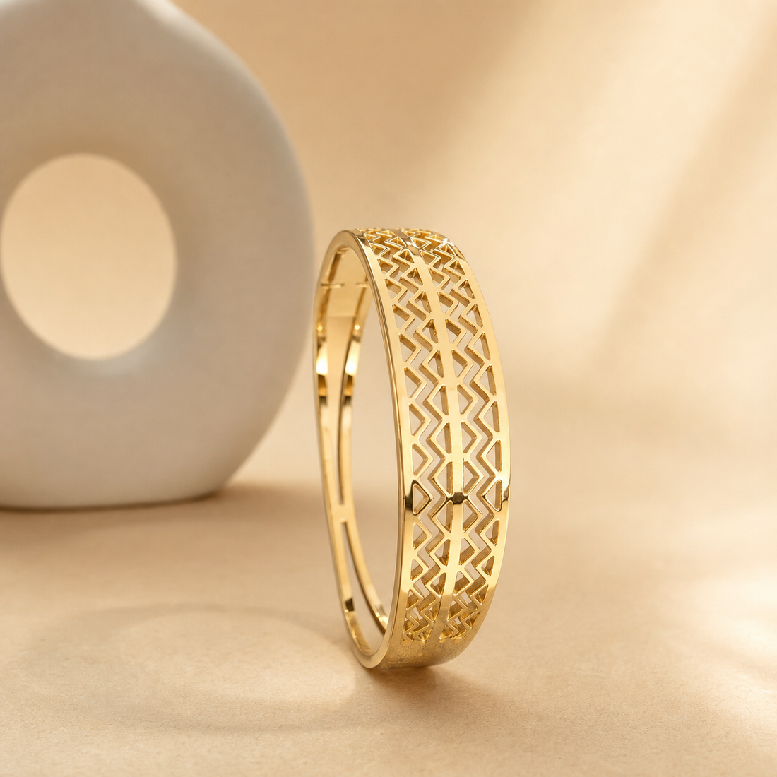 One rigid bangle in polished yellow gold. The band is roughly one-tenth of the outer diameter in width with a slightly rounded cross-section, and the width swells gently toward a broader face on one side. It has a hinged opening with a concealed box catch; the seam is visible but the hinge area is partly obscured. No stones, studs or applied motifs are present.