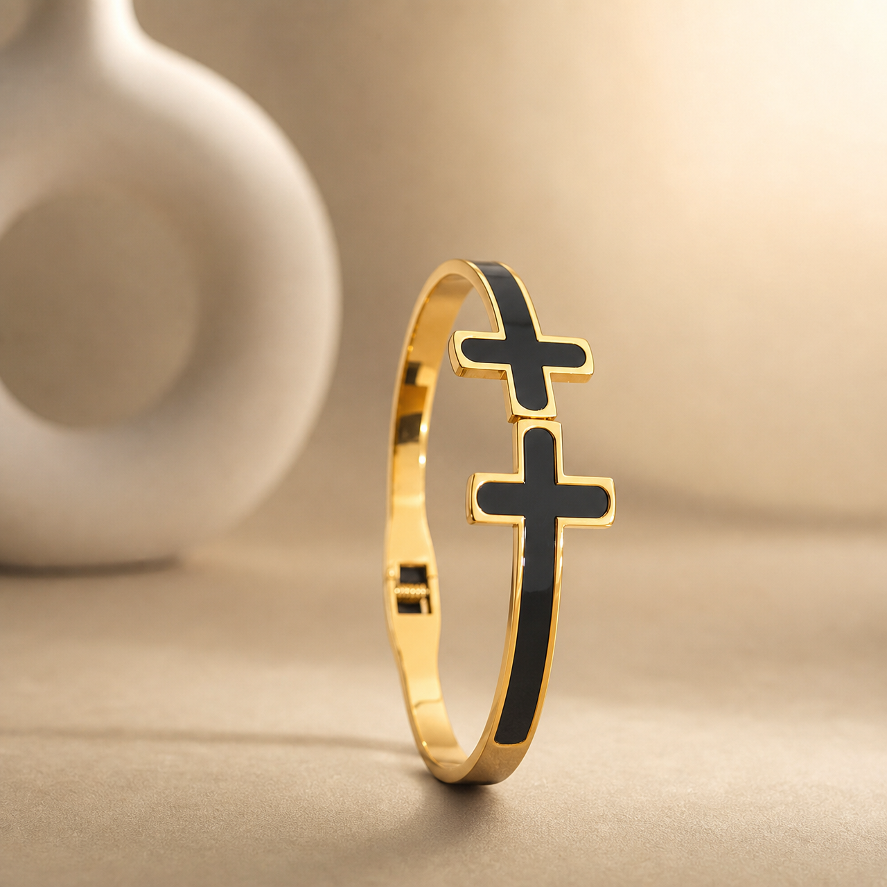 One rigid gold-tone metal bangle with a polished finish. The band is narrow, roughly one-twelfth of the outer diameter in width, of flat cross-section and even width all the way round, each face inlaid flush with black enamel panels running the full circumference. It is a hinged bangle with a box-style clasp opposite the hinge. Two solid-metal cross motifs with rounded arm ends, gold-rimmed and filled with black enamel, sit stacked on the band; the lower cross is larger than the one above it.