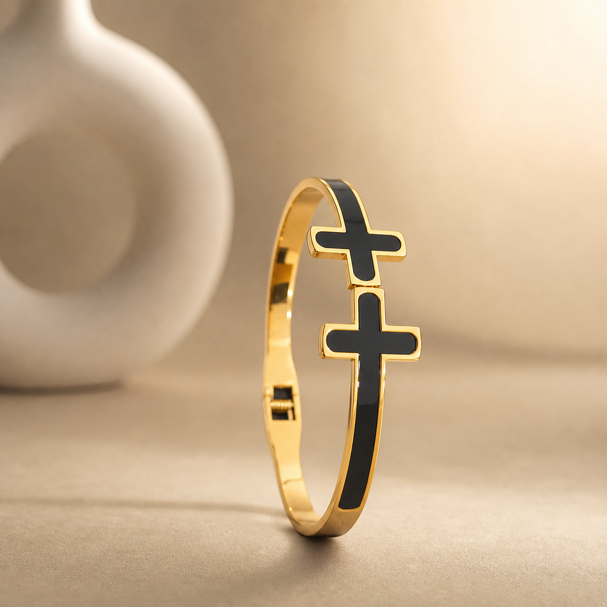 One rigid gold-tone metal bangle with a polished finish. The band is narrow, roughly one-twelfth of the outer diameter in width, of flat cross-section and even width all the way round, each face inlaid flush with black enamel panels running the full circumference. It is a hinged bangle with a box-style clasp opposite the hinge. Two solid-metal cross motifs with rounded arm ends, gold-rimmed and filled with black enamel, sit stacked on the band; the lower cross is larger than the one above it.