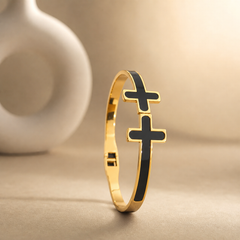 One rigid gold-tone metal bangle with a polished finish. The band is narrow, roughly one-twelfth of the outer diameter in width, of flat cross-section and even width all the way round, each face inlaid flush with black enamel panels running the full circumference. It is a hinged bangle with a box-style clasp opposite the hinge. Two solid-metal cross motifs with rounded arm ends, gold-rimmed and filled with black enamel, sit stacked on the band; the lower cross is larger than the one above it.