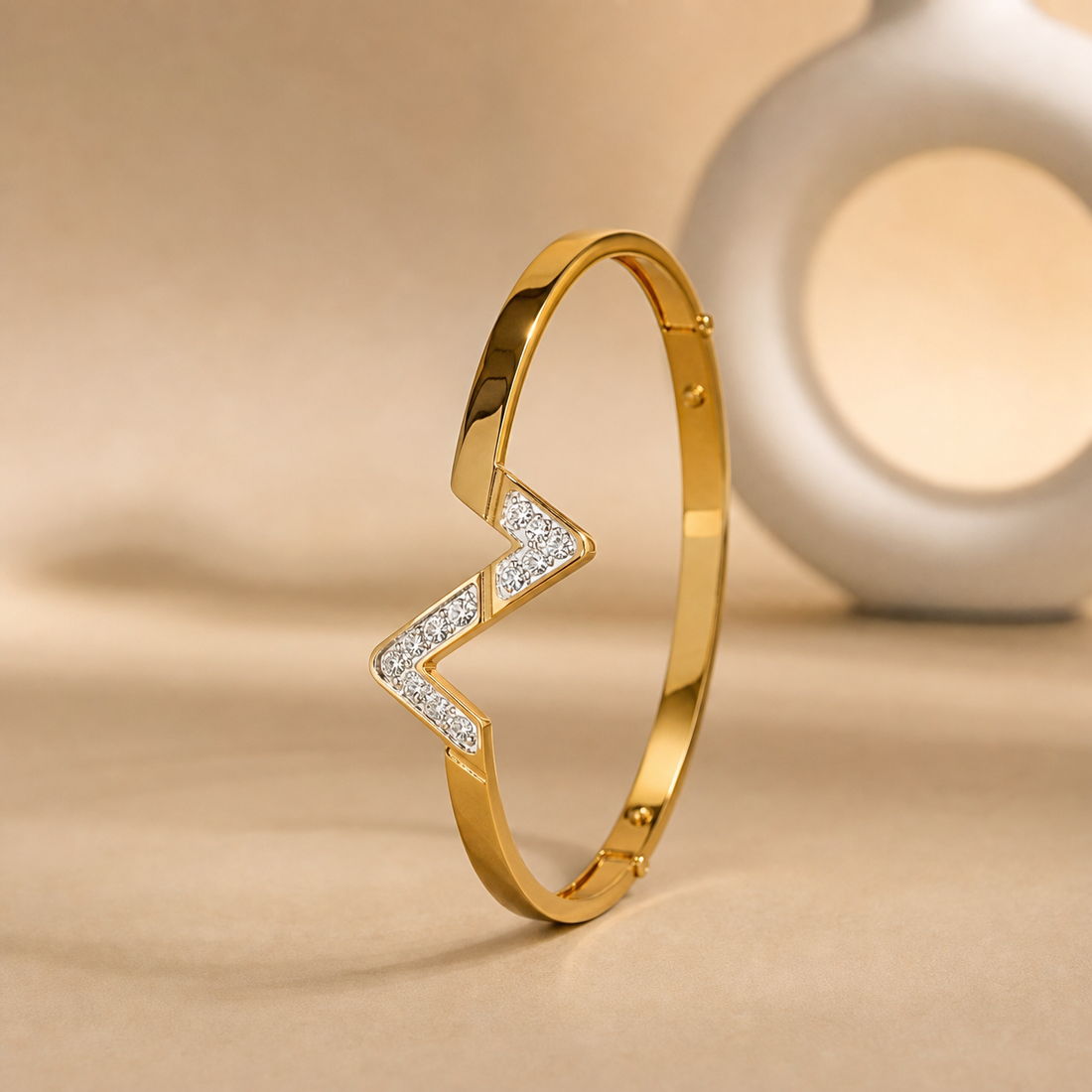 One rigid gold-tone bangle with a polished, mirror-finished yellow-metal surface. The band is flat in cross-section, its width roughly one-tenth of the outer diameter and its thickness slim, staying even most of the way round but interrupted by a broader applied V-shaped centrepiece. It is a fully closed rigid circle with small circular stud-like fixings visible at intervals along the band; the rear section is partly obscured.