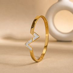 One rigid gold-tone bangle with a polished, mirror-finished yellow-metal surface. The band is flat in cross-section, its width roughly one-tenth of the outer diameter and its thickness slim, staying even most of the way round but interrupted by a broader applied V-shaped centrepiece. It is a fully closed rigid circle with small circular stud-like fixings visible at intervals along the band; the rear section is partly obscured.