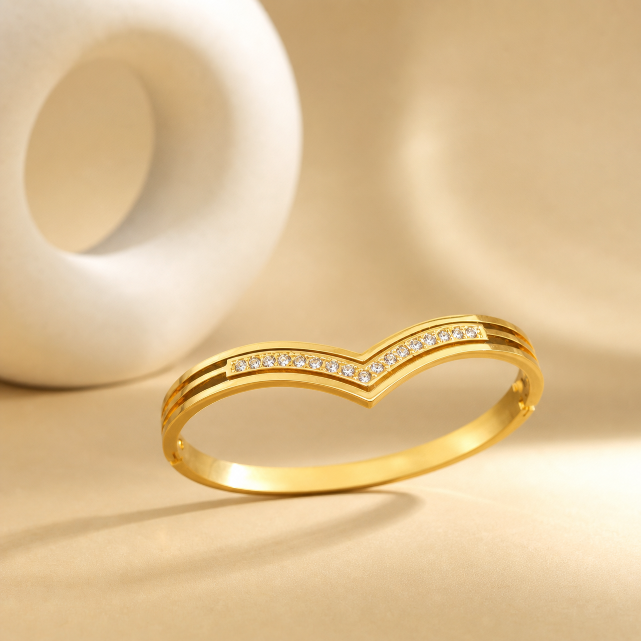 A single rigid gold-tone bangle with a bright high-polish finish. The band is narrow, roughly one-twentieth of the outer diameter in width, with a flat rectangular cross-section of even width all round except where the outer edge dips to a V-point on the face. It forms a fully closed oval circle with a screw fitting, small screw heads visible on the band.
