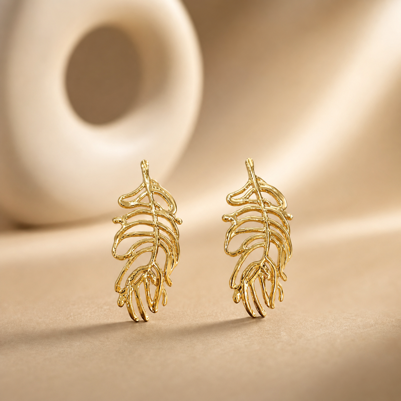 The photograph contains not a kada or bangle but a matched pair of two rigid gold-toned metal ear ornaments, each an openwork feather or leaf motif; no circular band, closure, hinge, gap, terminals or wrist-worn form is present, so band width, cross-section and closure type cannot be reported.