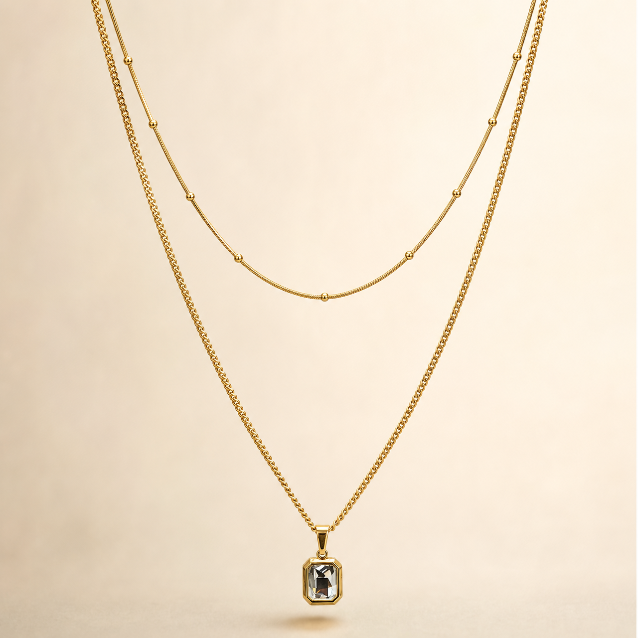 A two-strand necklace set in yellow-gold-tone metal with a polished finish. The set comprises two parallel strands joined at shared ends: a shorter, fine snake-style chain carrying small spherical bead stations spaced at irregular intervals along its run, and a longer, fine-gauge curb link chain from which a single pendant hangs. The longest chain run measures roughly 40 times the width of the pendant, the largest component; the shorter strand is approximately three-quarters the length of the long chain.