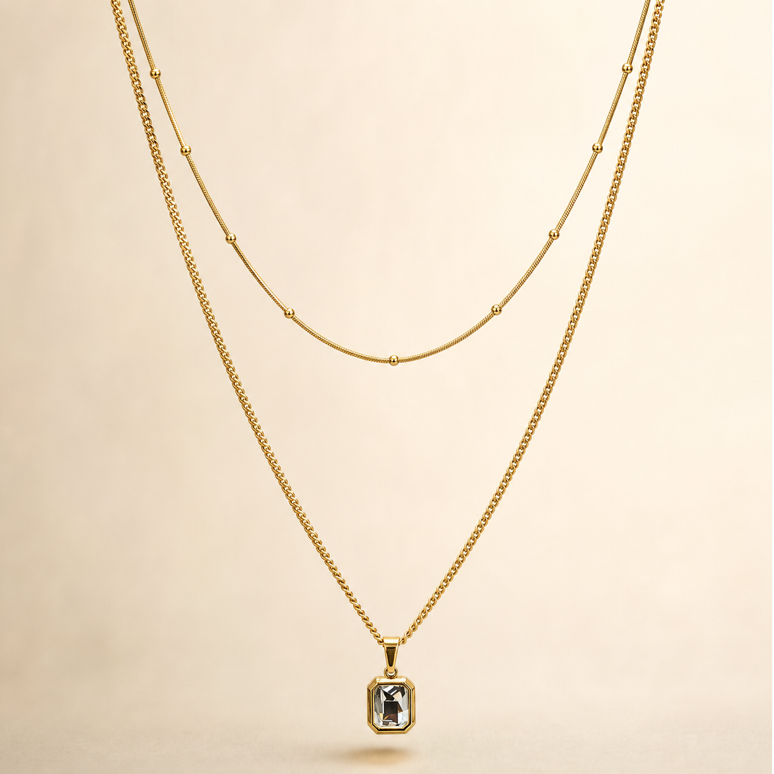 A two-strand necklace set in yellow-gold-tone metal with a polished finish. The set comprises two parallel strands joined at shared ends: a shorter, fine snake-style chain carrying small spherical bead stations spaced at irregular intervals along its run, and a longer, fine-gauge curb link chain from which a single pendant hangs. The longest chain run measures roughly 40 times the width of the pendant, the largest component; the shorter strand is approximately three-quarters the length of the long chain.