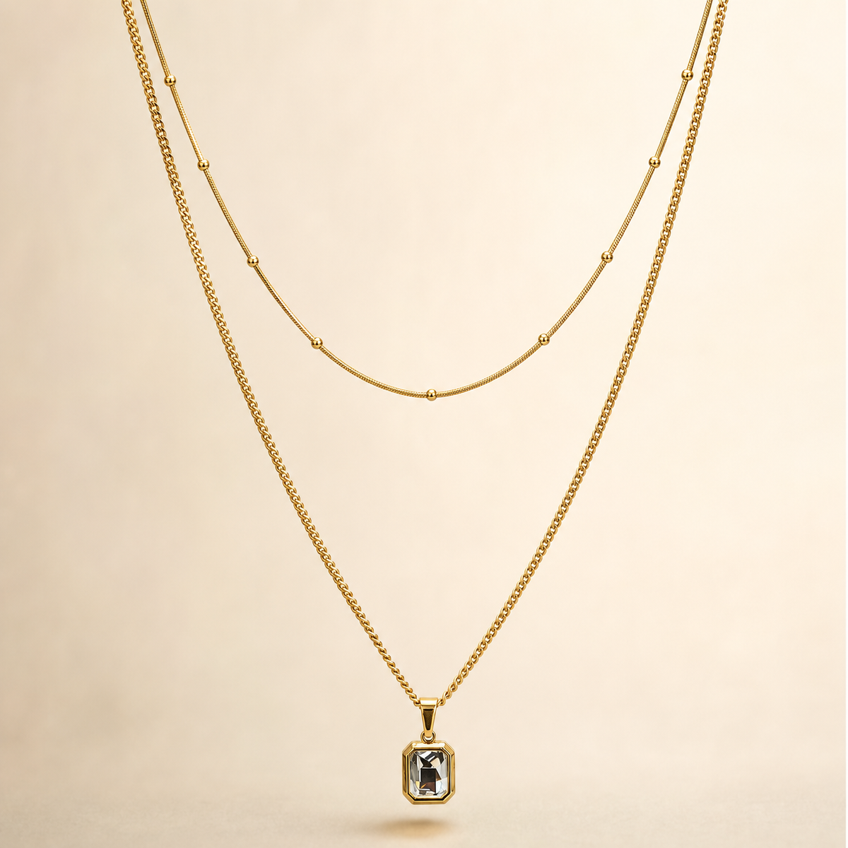 A two-strand necklace set in yellow-gold-tone metal with a polished finish. The set comprises two parallel strands joined at shared ends: a shorter, fine snake-style chain carrying small spherical bead stations spaced at irregular intervals along its run, and a longer, fine-gauge curb link chain from which a single pendant hangs. The longest chain run measures roughly 40 times the width of the pendant, the largest component; the shorter strand is approximately three-quarters the length of the long chain.
