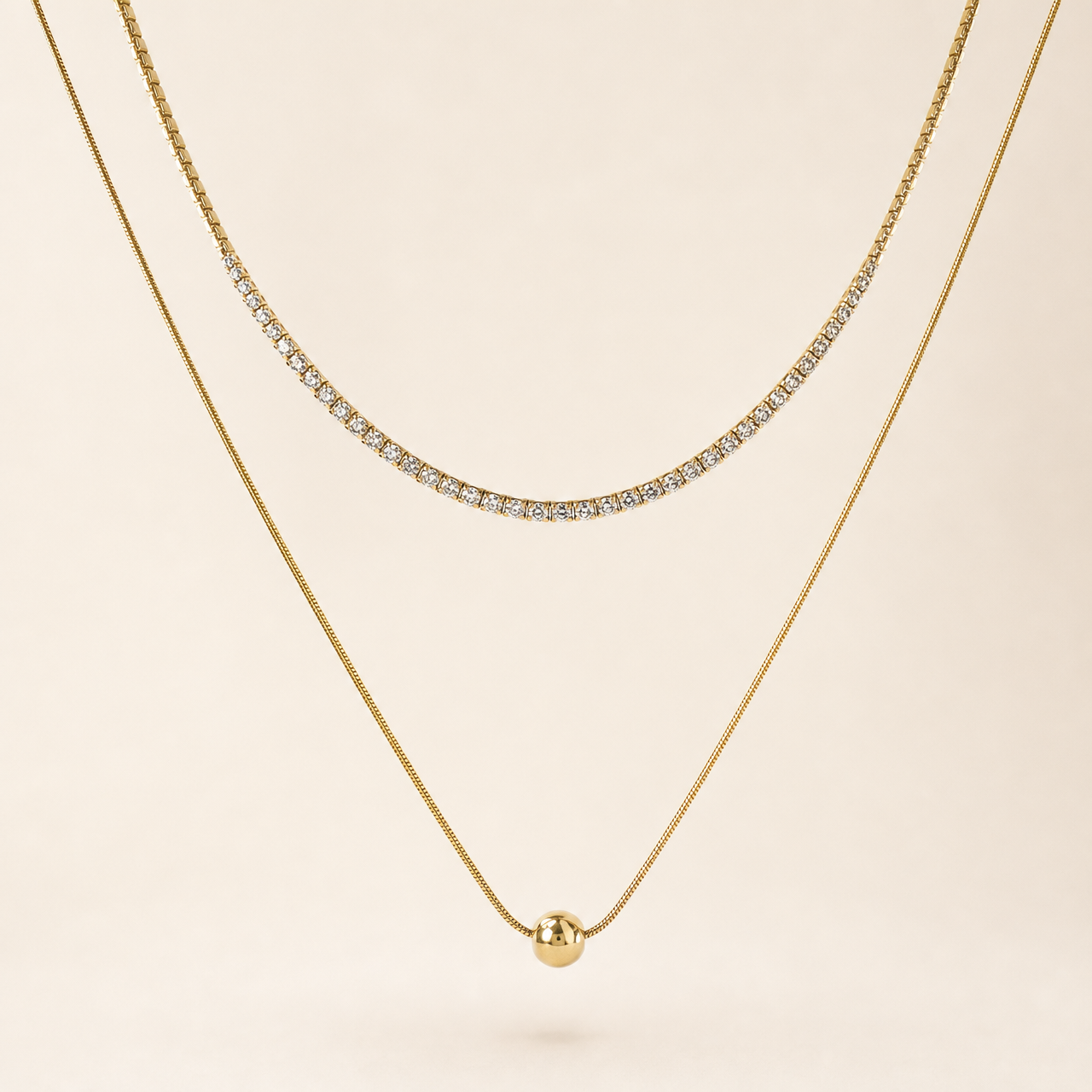 Two separate gold-tone necklaces, polished finish. Necklace one is a single-strand tennis-style chain of small clear round faceted stones, each in an individual gold box/prong mounting linked in a continuous articulated line; stone count is not clearly resolvable at the clasp ends and is withheld, stones are uniform and tightly spaced with no gaps; chain run is roughly 60-70 times one stone width, no extender visible.