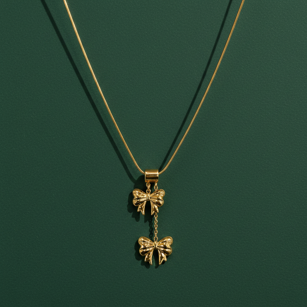 A single yellow-gold-tone necklace with a polished finish. Construction is one fine snake chain of small gauge forming a single strand, carrying a stacked double-bow pendant assembly: a smooth cylindrical barrel bail slides on the chain and supports, by short jump-ring links, a first solid-metal bow charm, from which a short fine chain drop suspends a second bow charm below it.