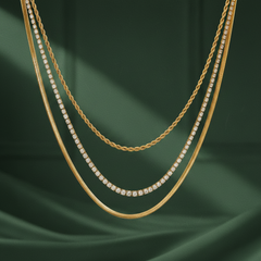 A layered set of three gold-tone metal chains with a polished finish, worn together as a multi-strand necklace. The strands are distinct in construction: a twisted rope chain of medium gauge; a tennis-style strand of individually claw-set clear round faceted stones in gold-tone box mounts, the stones graduating slightly larger toward the strand's middle; and a flat, flexible herringbone chain, the widest of the three.