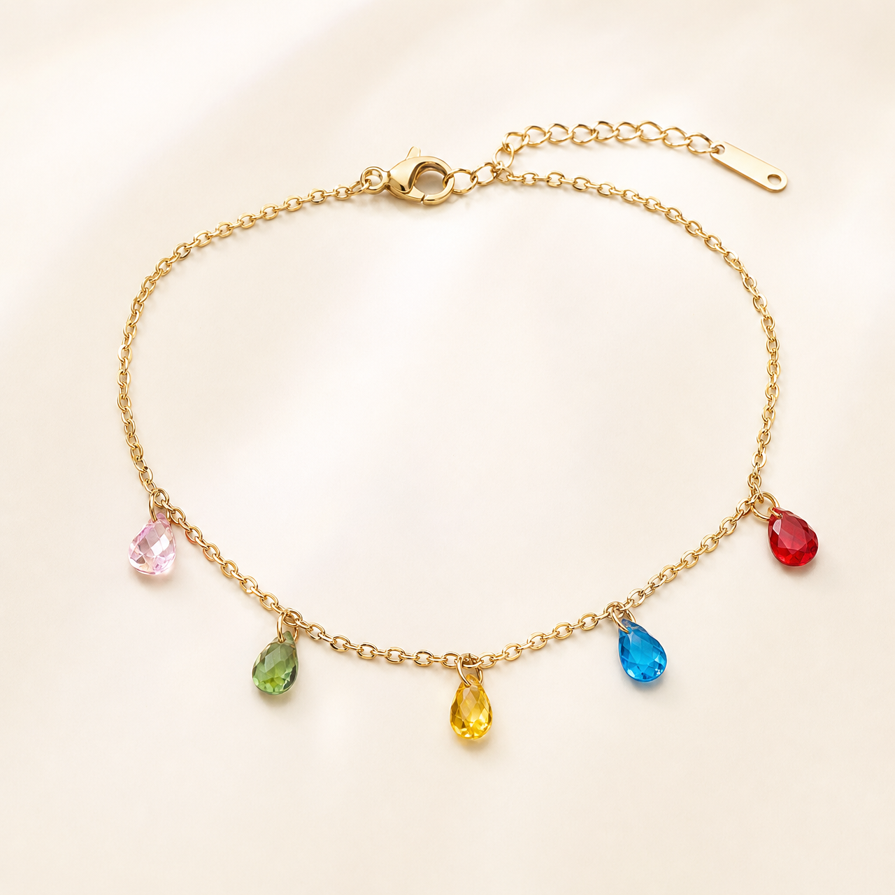 One anklet in warm gold-toned metal with a bright polished finish. It is a single strand of fine cable chain with small oval links of light gauge, closed by a working lobster-claw clasp. The main chain run is roughly thirty times the width of the largest charm, and a short extender chain of larger oval links, about one fifth the length of the main chain, ends in a single elongated flat terminal tag.