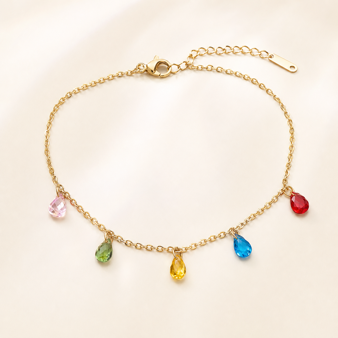 One anklet in warm gold-toned metal with a bright polished finish. It is a single strand of fine cable chain with small oval links of light gauge, closed by a working lobster-claw clasp. The main chain run is roughly thirty times the width of the largest charm, and a short extender chain of larger oval links, about one fifth the length of the main chain, ends in a single elongated flat terminal tag.