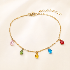 One anklet in warm gold-toned metal with a bright polished finish. It is a single strand of fine cable chain with small oval links of light gauge, closed by a working lobster-claw clasp. The main chain run is roughly thirty times the width of the largest charm, and a short extender chain of larger oval links, about one fifth the length of the main chain, ends in a single elongated flat terminal tag.