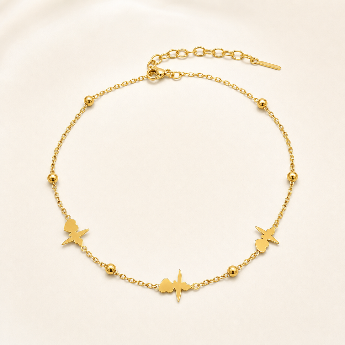 One anklet in yellow-gold-toned metal with a polished finish. It is a single flexible strand of fine, small-gauge oval cable chain with no branches. A lobster-claw clasp closes one end, connecting to a short extender of larger open oval links terminating in a plain straight bar tag; the extender is roughly one quarter of the main chain run, and the full chain run is approximately 25 to 30 times the width of the largest charm. A working closure is visible.