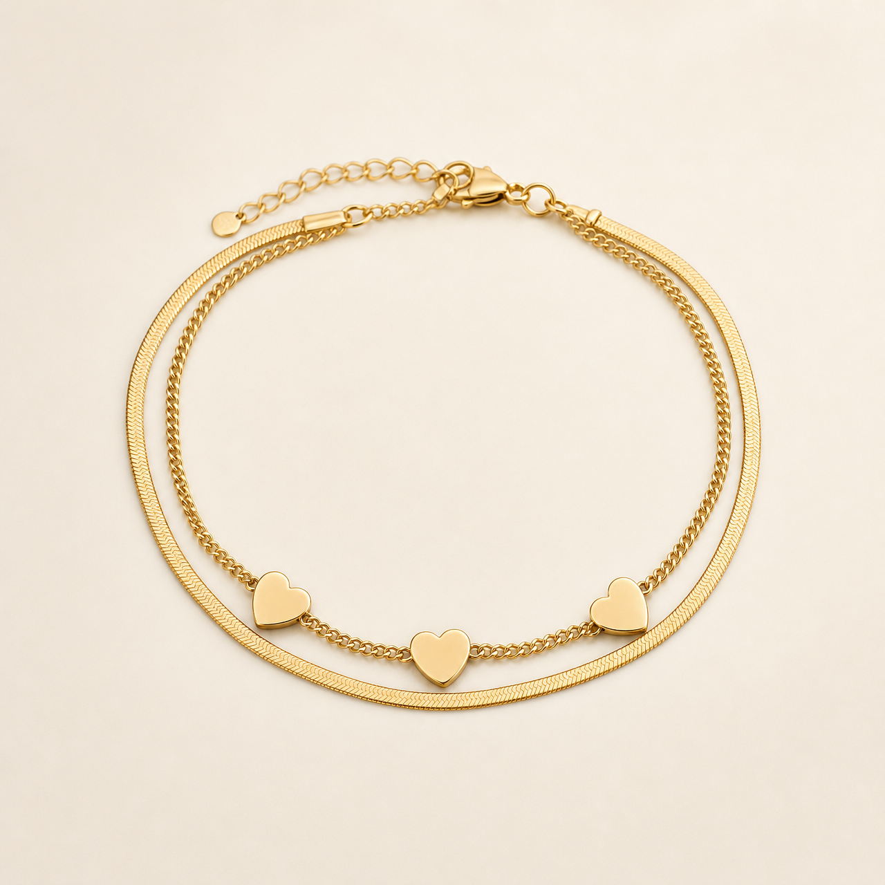 One anklet in yellow-gold-coloured metal with a polished finish. It is a double-strand flexible chain: one strand is a flat, smooth herringbone/snake chain of fine gauge, and the other is a finer rounded curb chain, the two strands joined together at both ends. The main chain run measures roughly twenty-five to thirty times the width of the largest component (the heart charm); a short extender chain of open links, about one eighth of the main chain length, runs from one end.