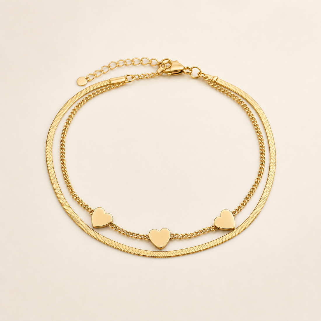 One anklet in yellow-gold-coloured metal with a polished finish. It is a double-strand flexible chain: one strand is a flat, smooth herringbone/snake chain of fine gauge, and the other is a finer rounded curb chain, the two strands joined together at both ends. The main chain run measures roughly twenty-five to thirty times the width of the largest component (the heart charm); a short extender chain of open links, about one eighth of the main chain length, runs from one end.