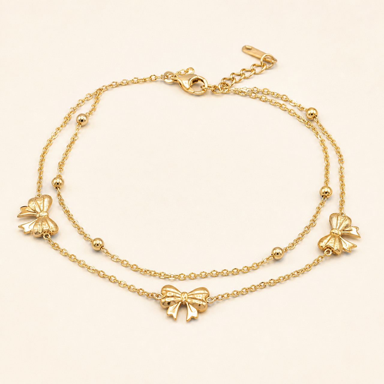 One anklet: a gold-toned polished flexible ankle chain in a two-strand layered build, both fine oval-link cable chains of small gauge joining at a common clasp ring and continuing as connected decorated runs. The main chain run is approximately twelve to fourteen times the width of the largest bow motif; the extender is about one-fifth of the main chain and ends in one flat terminal tag. A working lobster clasp is visible.