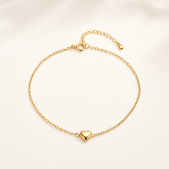 One anklet in yellow-gold-toned metal with a polished finish. It is a single-strand, fine-gauge cable chain of small round links. The chain run is approximately 45 to 50 times the width of its largest component, the heart charm; the extender chain is roughly one-sixth the length of the main chain and ends in a single small elongated teardrop terminal tag. A working spring-ring clasp is visible, connecting to a jump ring.