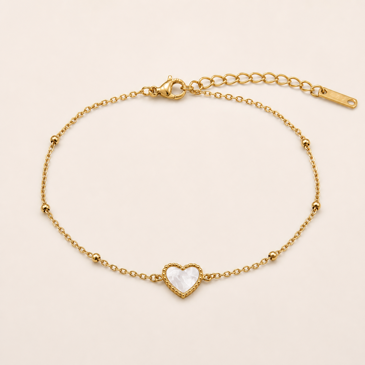 One anklet in yellow-gold-tone metal with a polished finish. It is a single fine cable chain of small oval links, one continuous strand. From the clasp end: a lobster clasp, working and visible, attaches to a short section of chain; a long extender chain of larger curb-style links, roughly one quarter the main chain's length, ends in a flat rectangular terminal tag, one tag total. The main chain run is approximately 20 times the width of the largest component, the heart charm.