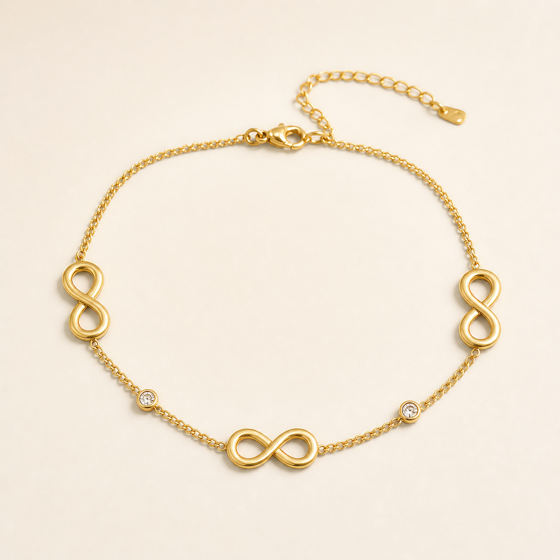 One anklet in polished yellow gold-tone metal, a single flexible strand of fine curb chain of small, even links. From the lobster-claw clasp end the strand runs as plain chain before meeting a solid-metal infinity-symbol station, then a small round clear faceted stone in a flush bezel setting, then a second infinity station, then a second bezel-set clear round stone, then a third infinity station, after which plain chain runs to the jump-ring terminal; decoration thus alternates motif and stone around the