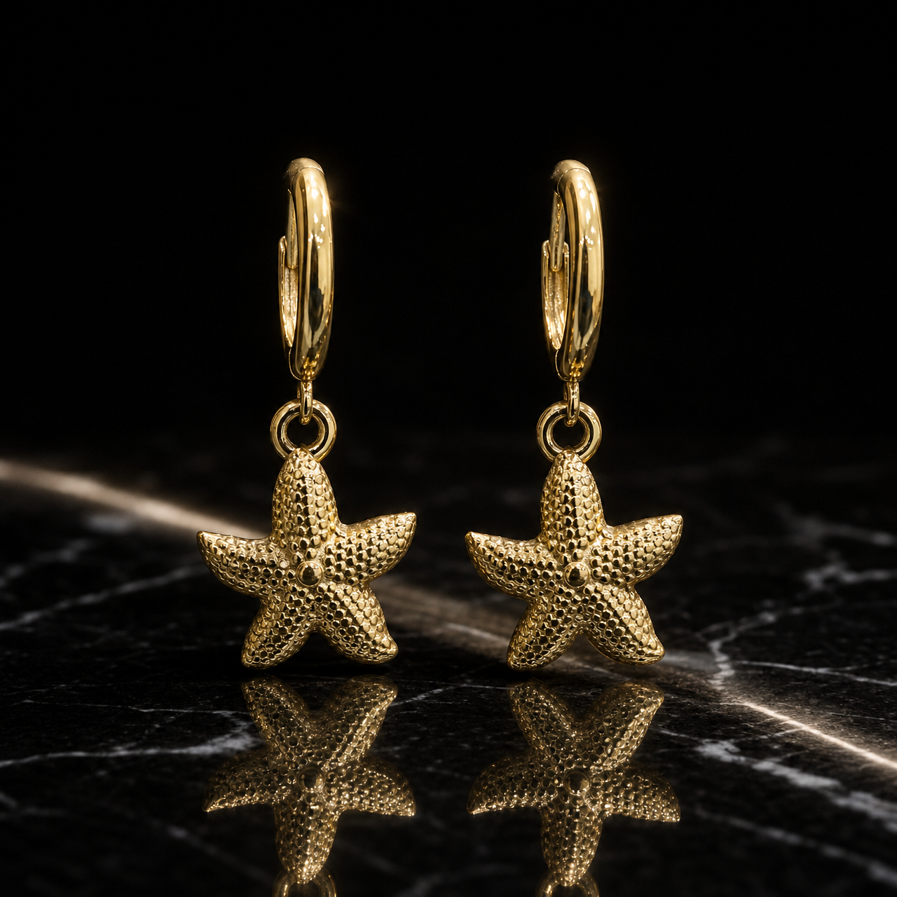 Two earrings, one design: yellow-gold-toned metal with polished fittings and a granular hammered texture on the motifs. Each earring has a small huggie-hinge hoop of medium gauge, with overall drop approximately twice the widest starfish-arm span. From each hoop a single jump ring mounts one solid five-arm starfish dangle, textured across its surface with a small raised central dot, swinging freely at the jump ring. No stones, pearls, discs or tassels are present.