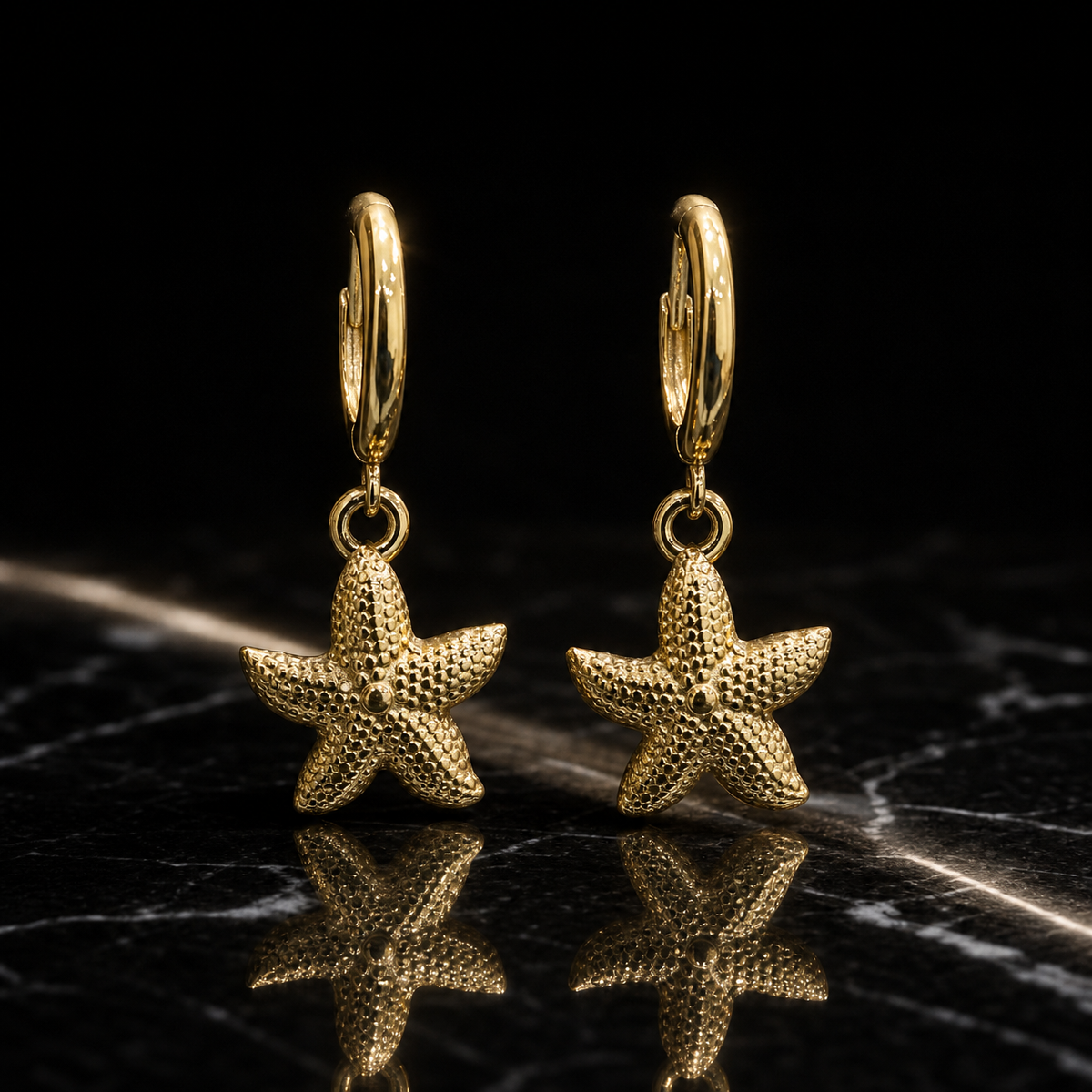 Two earrings, one design: yellow-gold-toned metal with polished fittings and a granular hammered texture on the motifs. Each earring has a small huggie-hinge hoop of medium gauge, with overall drop approximately twice the widest starfish-arm span. From each hoop a single jump ring mounts one solid five-arm starfish dangle, textured across its surface with a small raised central dot, swinging freely at the jump ring. No stones, pearls, discs or tassels are present.