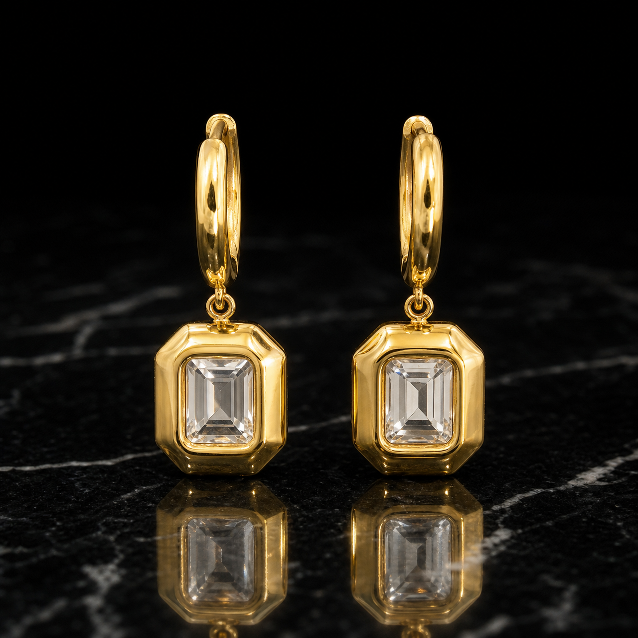 Two earrings of one design in yellow-gold-tone metal with a polished finish. Each has a huggie hinge hoop fitting of short length and medium gauge, with an overall drop about twice the pendant's widest part. From the hoop, a small jump ring articulates a single rigid section: an octagonal rectangular frame pendant. Set within each frame is one clear rectangular step-cut faceted element in a bezel mounting, centred in a solid polished border.