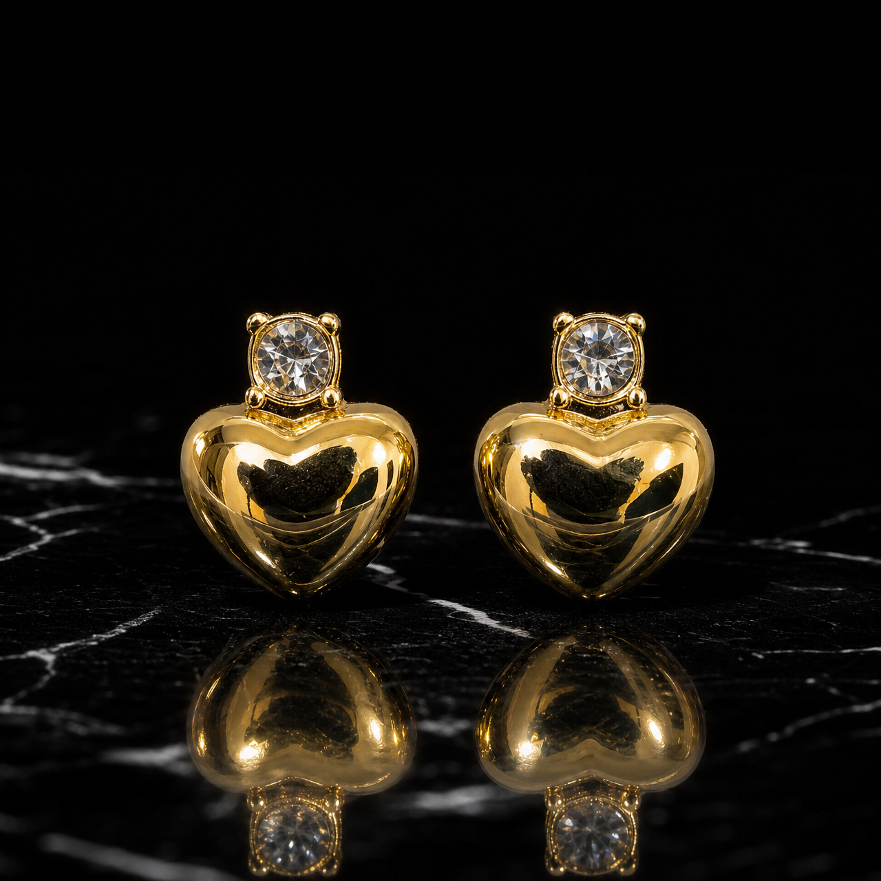 Two earrings. Yellow-gold-coloured metal with a high-polish, mirror finish on the main motif. Each is an ear-worn stud with a short post-and-back fitting of fine gauge. The overall drop is roughly one-and-a-half to two times the widest part (the heart). Each has two sections: a stud head and a single drop that swings slightly beneath it. From the fitting downward: one small colourless round faceted element in a four-prong crown mount; directly below, one solid puffed heart forming the polished drop.