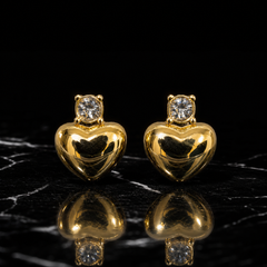 Two earrings. Yellow-gold-coloured metal with a high-polish, mirror finish on the main motif. Each is an ear-worn stud with a short post-and-back fitting of fine gauge. The overall drop is roughly one-and-a-half to two times the widest part (the heart). Each has two sections: a stud head and a single drop that swings slightly beneath it. From the fitting downward: one small colourless round faceted element in a four-prong crown mount; directly below, one solid puffed heart forming the polished drop.