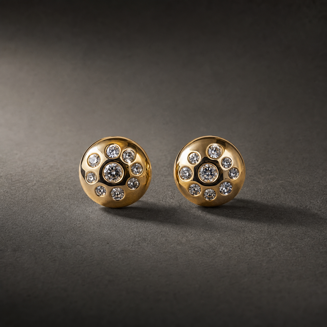 Two ear-worn stud earrings of one design. Each is a polished yellow-gold domed disc, round in outline, its surface flush-set with clear round faceted stones: one larger central stone surrounded by smaller graduated stones scattered across the dome, all in smooth bezel-style recessed settings; exact stone count is not fully resolvable. The earrings are rigid, single-section studs with no articulation or dangling parts; overall depth is roughly equal to the disc width.
