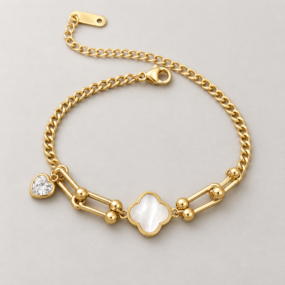 Gold-tone 304 stainless steel chain bracelet with a mother-of-pearl clover charm and a clear heart-cut stone drop, laid flat on a grey surface