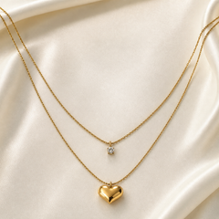 A two-strand layered pendant necklace in yellow-gold-coloured metal with a polished, reflective finish. Both strands are fine twisted rope-style chains of similar gauge, running roughly parallel and diverging only in length. The shorter, inner strand carries one pendant: a single round clear faceted stone, approximately 4 to 5 mm, held in a multi-prong setting suspended from a small jump-ring bail at the chain's centre.