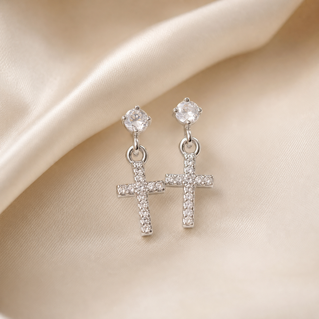 Two matching drop earrings, a pair. Silver-coloured metal with a bright polished finish. Each earring is built from an ear stud carrying one round clear faceted element in a raised claw/prong mounting, linked by one small oval metal jump ring to a dangling Latin-cross pendant; the post and any back fitting are partly obscured by the fingers and are not counted.