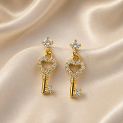 A matched pair of two dangle stud earrings in yellow-gold-toned metal with a polished finish. Each earring consists of a single round clear faceted element in a four-prong setting at the top, forming the stud, from which a key-shaped pendant hangs by two small interlocked jump rings, allowing the pendant to dangle.