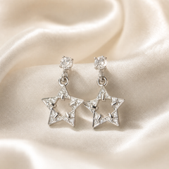 A matched pair of 2 drop stud earrings in silver-coloured metal with a polished bright finish. Each earring comprises 1 round clear faceted element held in a multi-prong stud setting at the ear, connected by a small round jump ring to a dangling five-pointed open star motif of solid metal with a hollow centre.