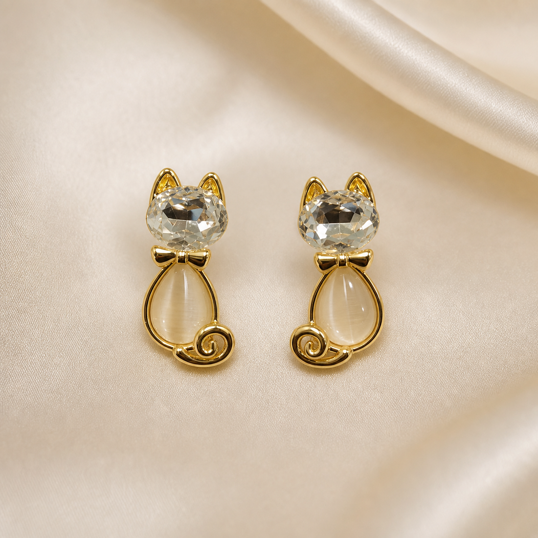 A matched pair of cat-shaped stud earrings in polished yellow gold-tone metal. Each earring is a single cast motif of a seated cat: the head is a large, round, clear faceted element set in the metal, topped by two pointed solid-metal ears; below the head sits a small solid-metal bow-tie collar; the body is an elongated teardrop-shaped translucent cabochon, milky cream in colour with a soft internal sheen, enclosed in a thin gold wire frame that follows the teardrop outline and finishes in a curled spiral