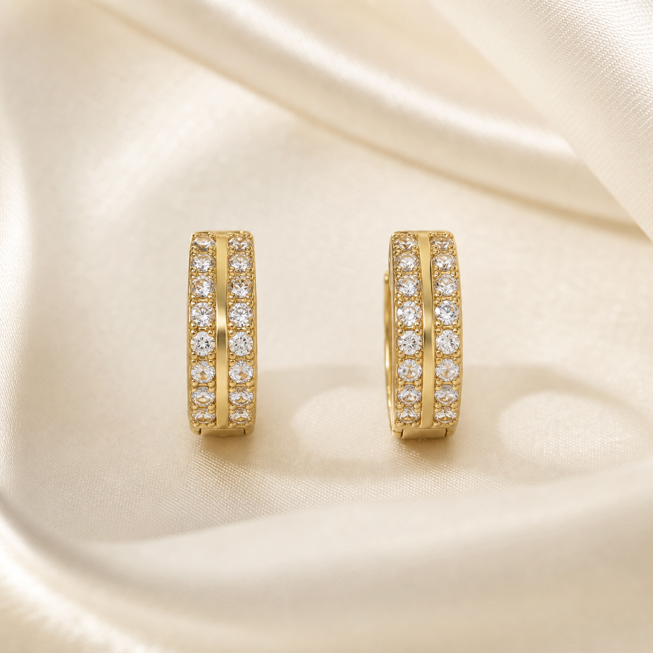 A matched pair of two small huggie-style hoop earrings in yellow-gold-tone metal with a polished finish. Each earring is a rigid, narrow oval hoop of consistent width, its outer face carrying two parallel rows of small, round, clear colourless faceted stones running the full length of the band; the rows are separated by a slim polished gold channel ridge.