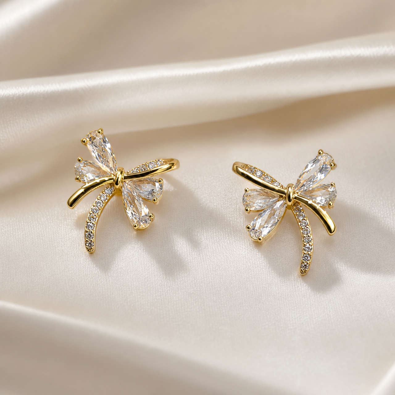 A matched pair of two bow-shaped stud earrings in gold-tone metal with a polished, bright finish. Each earring is a single cast bow motif with a small knotted centre from which four loops and two trailing ribbon ends radiate; there is no chain, strand, clasp or extender, the reverse fittings being partly obscured.