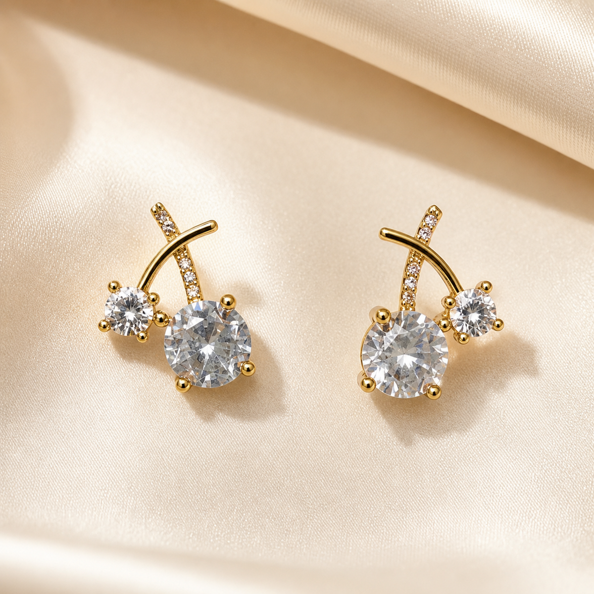 A matched pair of stud earrings, two items total, in yellow-gold-toned polished metal. Each earring is built around two crossing curved bar stems forming an X-like junction above the stones; one stem of each earring carries a line of small channel-set or pavé-set clear faceted accent stones along its length, while the crossing stem is plain polished metal.
