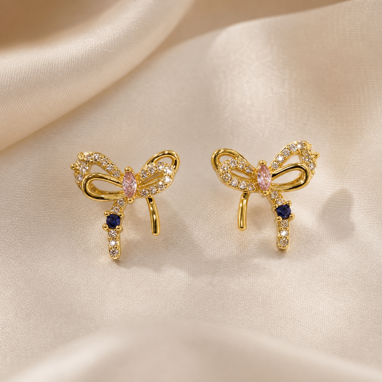 A matched pair of stud earrings, two items, each a solid-metal bow motif in yellow-gold tone with a polished finish on the smooth ribbon sections. Each bow is formed from a single cast openwork ribbon with two loops and two trailing ribbon ends, one tail plain polished metal and the other set with a continuous field of small round clear faceted stones in pavé. At the knot centre of each bow sits one marquise-cut pink faceted stone in a prong setting, its long axis vertical.