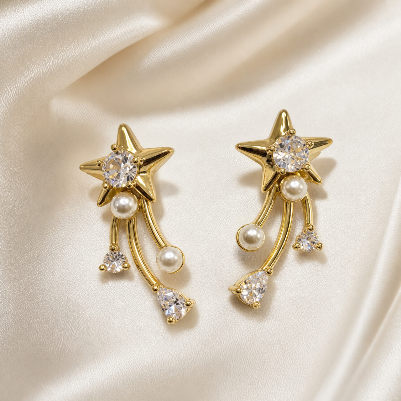 A matched pair of two drop earrings in polished yellow-gold-tone metal. Each earring has a stud-style top formed as a solid five-pointed star with domed, polished relief, its centre holding one large round brilliant-cut clear faceted stone in a prong setting. Beneath the star, one small white round faux pearl is fixed at the junction where three curved, slender gold-tone bar strands hang freely; the strands are of slightly differing lengths and each terminates in a dangling element.