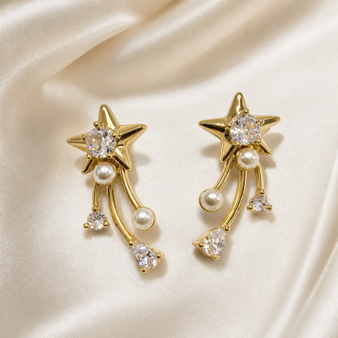 A matched pair of two drop earrings in polished yellow-gold-tone metal. Each earring has a stud-style top formed as a solid five-pointed star with domed, polished relief, its centre holding one large round brilliant-cut clear faceted stone in a prong setting. Beneath the star, one small white round faux pearl is fixed at the junction where three curved, slender gold-tone bar strands hang freely; the strands are of slightly differing lengths and each terminates in a dangling element.