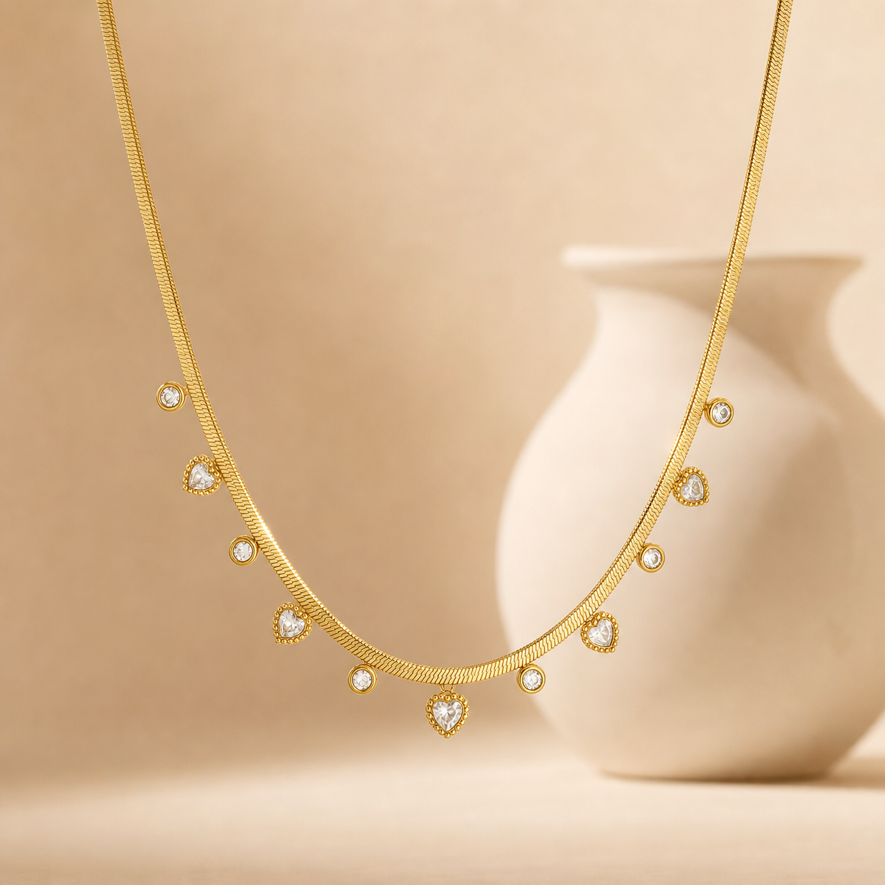 One gold-toned necklace of a single flat snake (herringbone-style) chain, medium gauge, with a smooth flexible ribbon-like surface and polished finish. The chain run is roughly 40 times the width of the largest heart charm; no extender is visible in view, so extender length is indeterminate (the ends and any clasp are partly obscured and not resolvable).